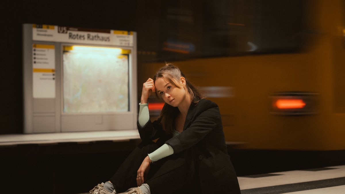 A young woman in casual attire seated at a Berlin subway station with trains in motion.